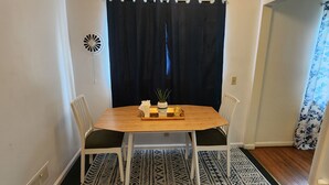 Un comedor con una mesa de madera, sillas blancas, una cortina negra y una alfombra estampada.