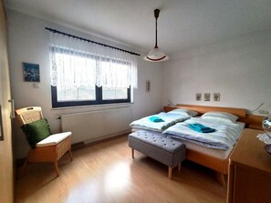 Een slaapkamer met een bed, een stoel, een raam met gordijnen, een radiator en een plafondlamp.