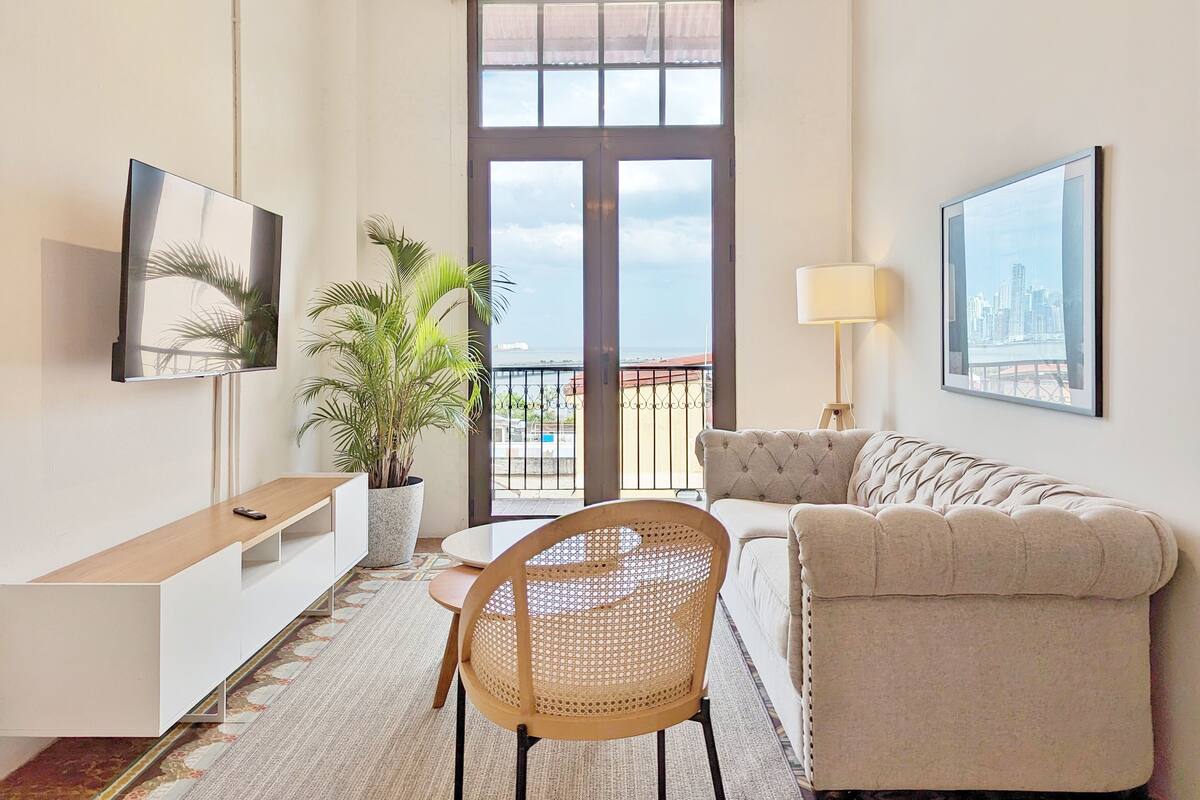 Uma sala de estar moderna com sofá, televisão, planta em vaso, mesa de jantar e vista para o mar através de uma grande janela.