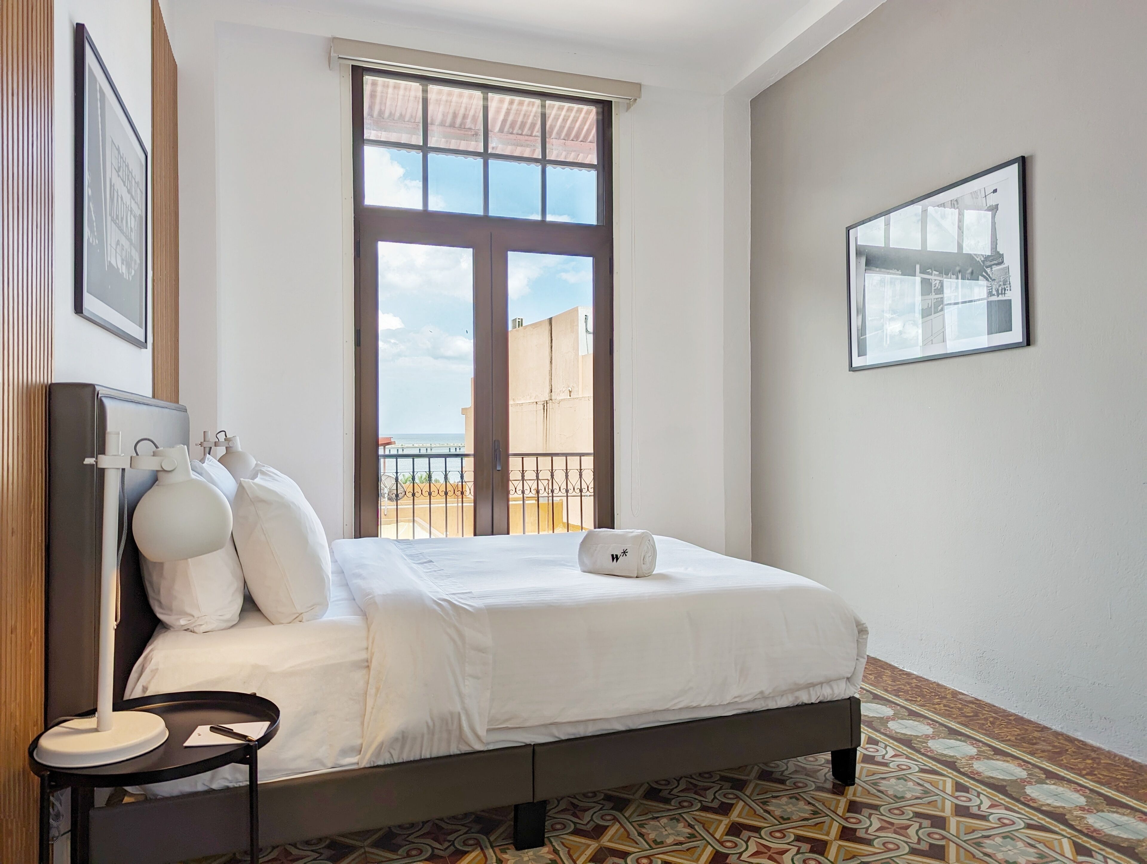 A neatly made bed with white linens, a bedside table with a lamp, and a large window with a view of buildings and a clear sky.