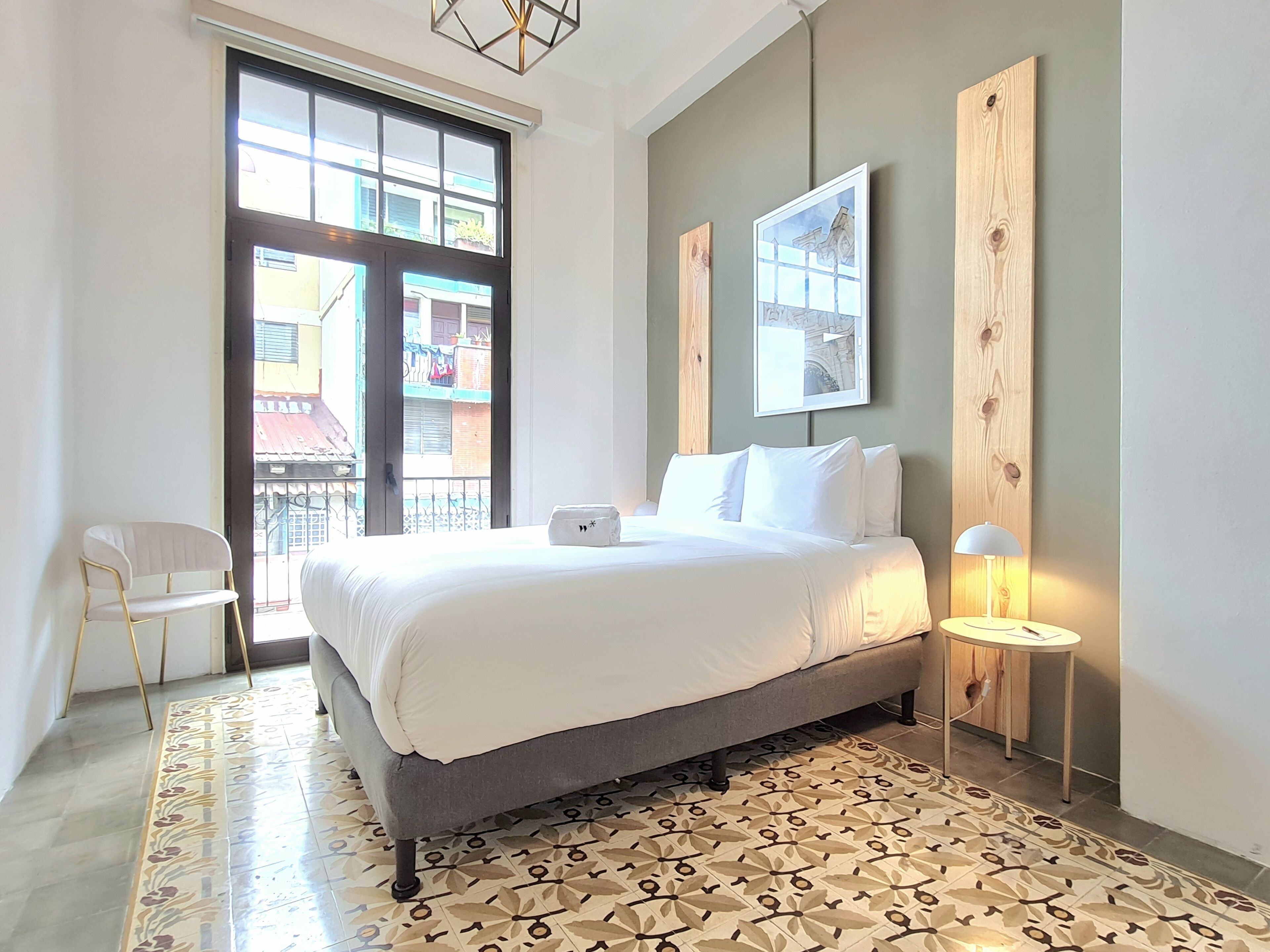 Una habitación de hotel moderna con una cama grande, una mesita auxiliar pequeña, una silla y vistas a través de un ventanal amplio.
