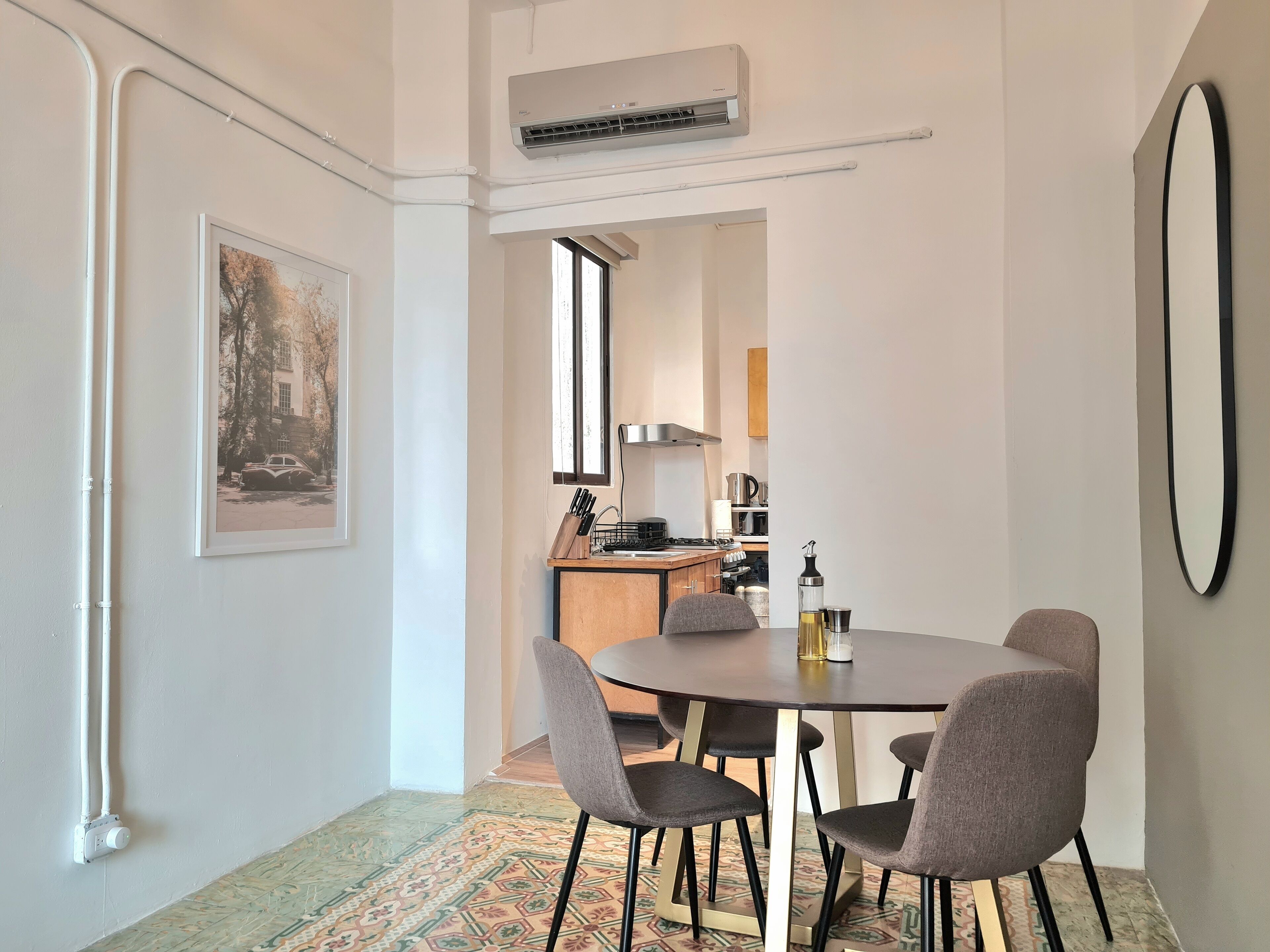 A modern dining area with a round table and four chairs, an air conditioning unit above, a framed picture on the wall, and a kitchen in the background.