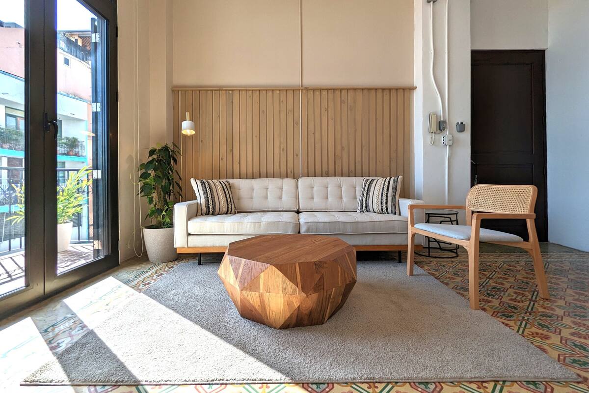 Uma sala de estar com sofá, mesa de centro de madeira, cadeira, planta em vaso e tapete estampado.