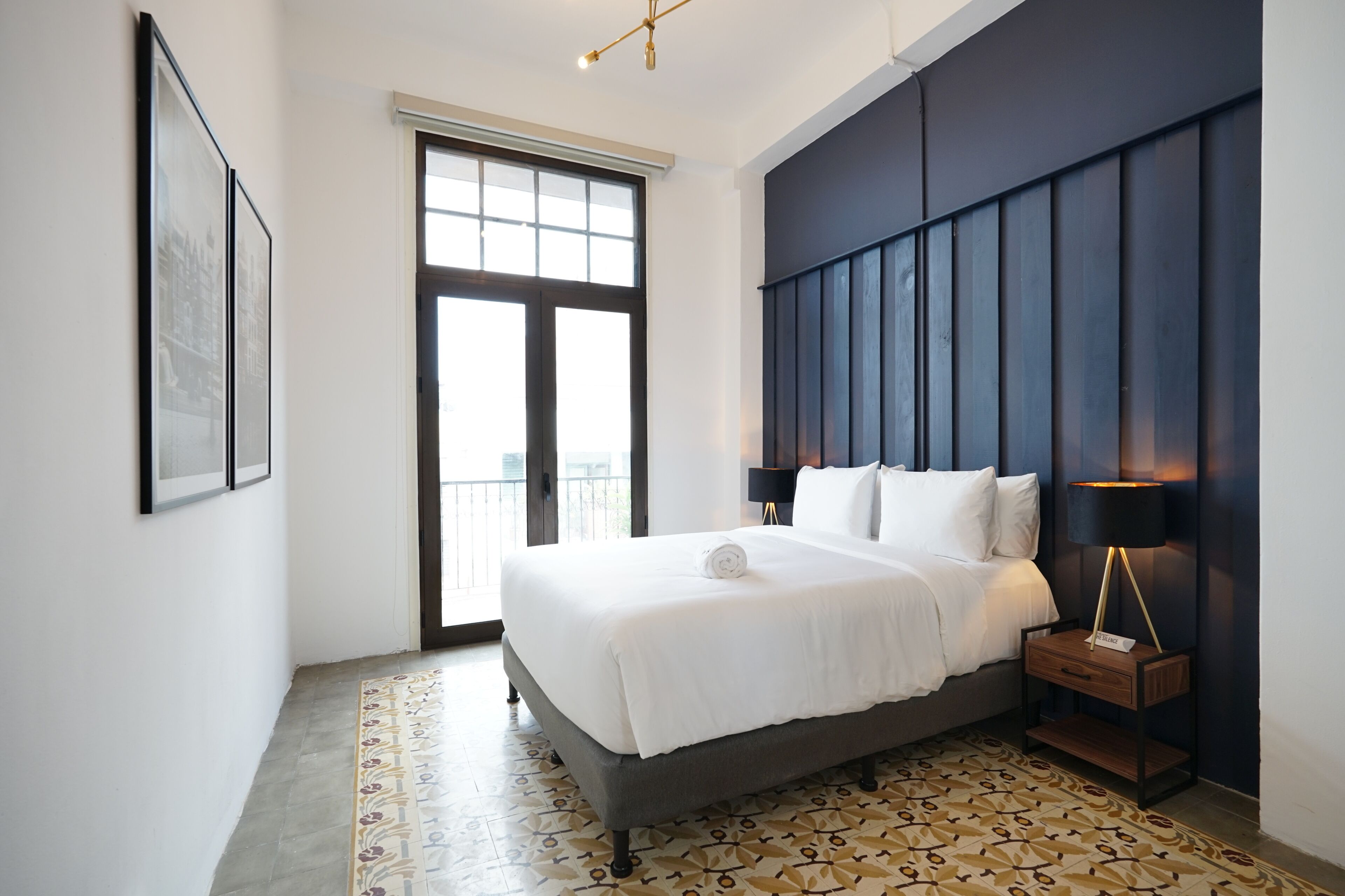 A modern hotel room with a large bed, a nightstand, a wall-mounted lamp, and a patterned floor.