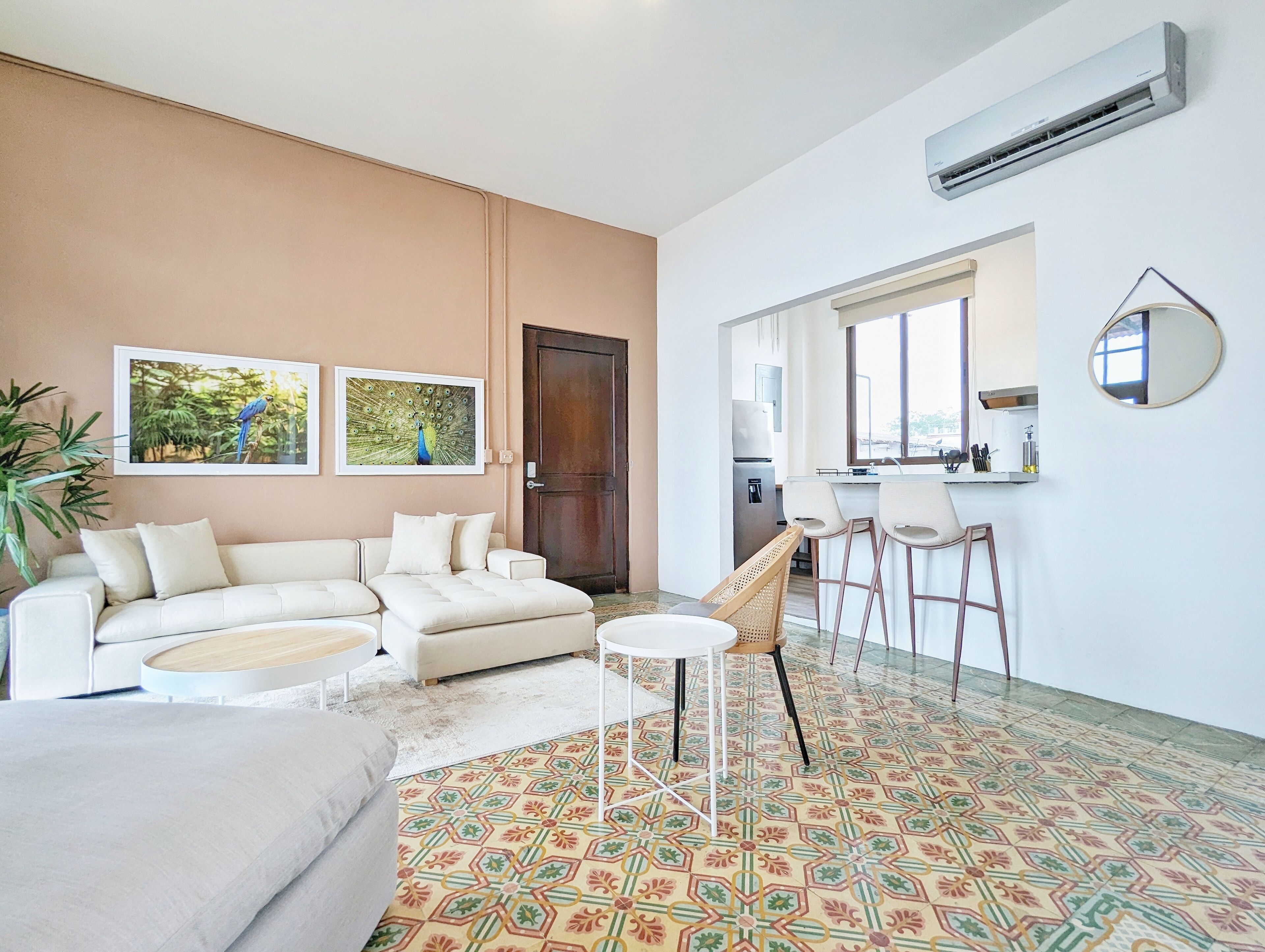 A modern living room with a sofa, a coffee table, and a patterned floor. There is a door leading to a kitchen area with a refrigerator and a window.