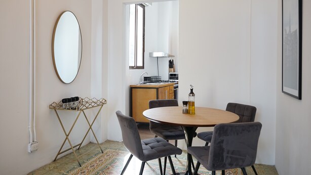 A modern dining area with a round table and six chairs, a small side table with a bottle and glasses, a large mirror, and a view into a kitchen area. - Donovan Night Scene by Wynwood (Panama City)