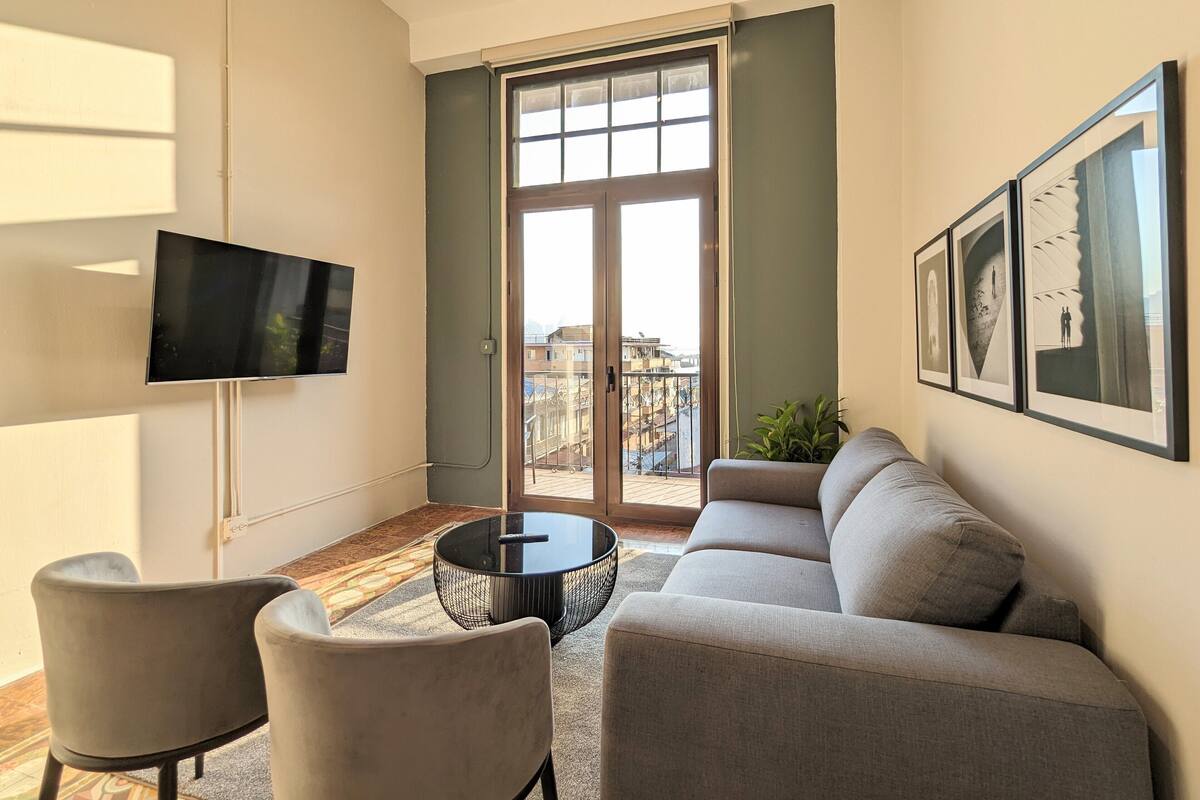 Uma sala de estar moderna com sofá, cadeiras, mesa de centro, televisão e varanda com vista para a cidade.