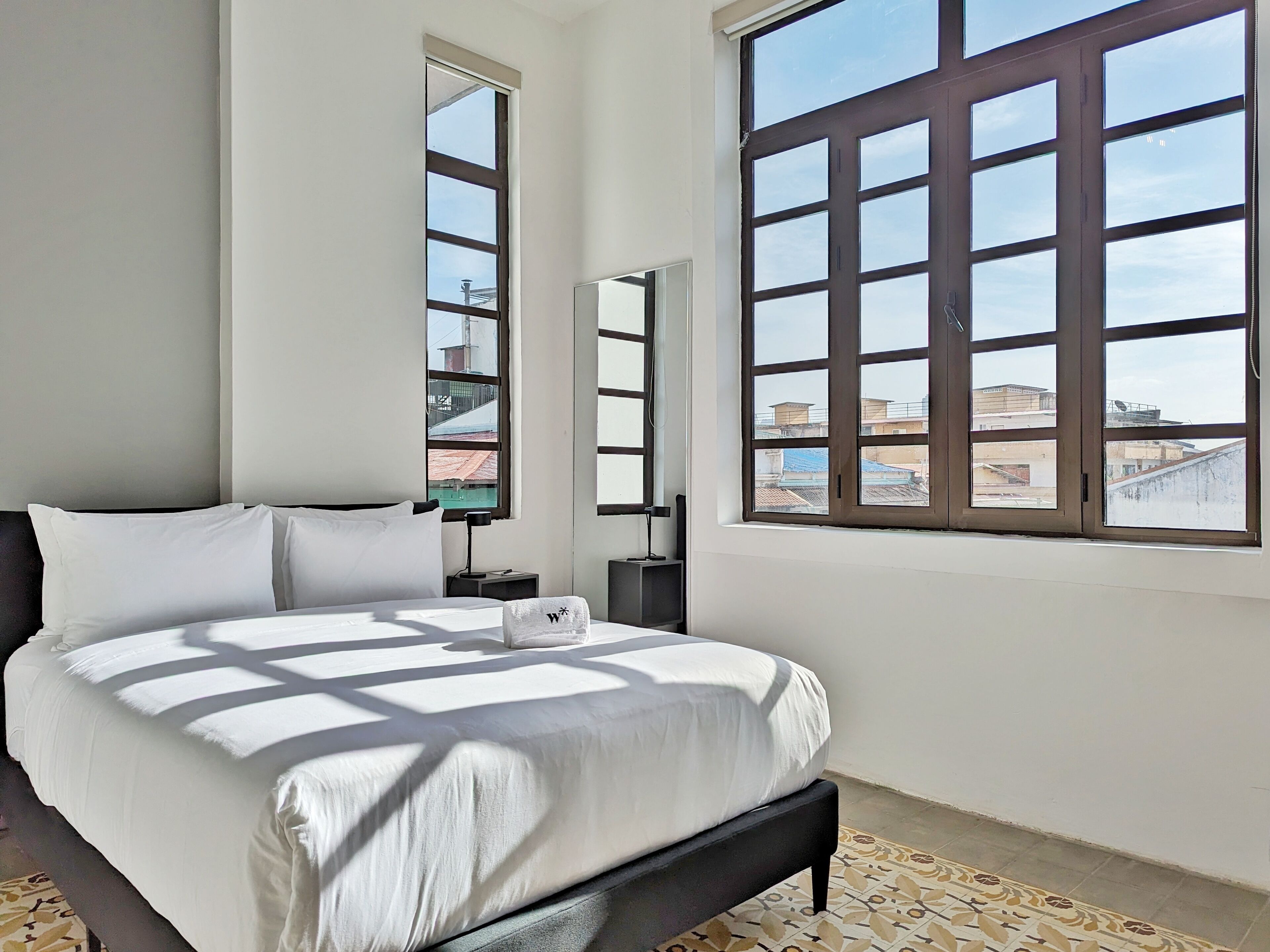 A bedroom with a large bed, two windows, a nightstand, and a mirror.
