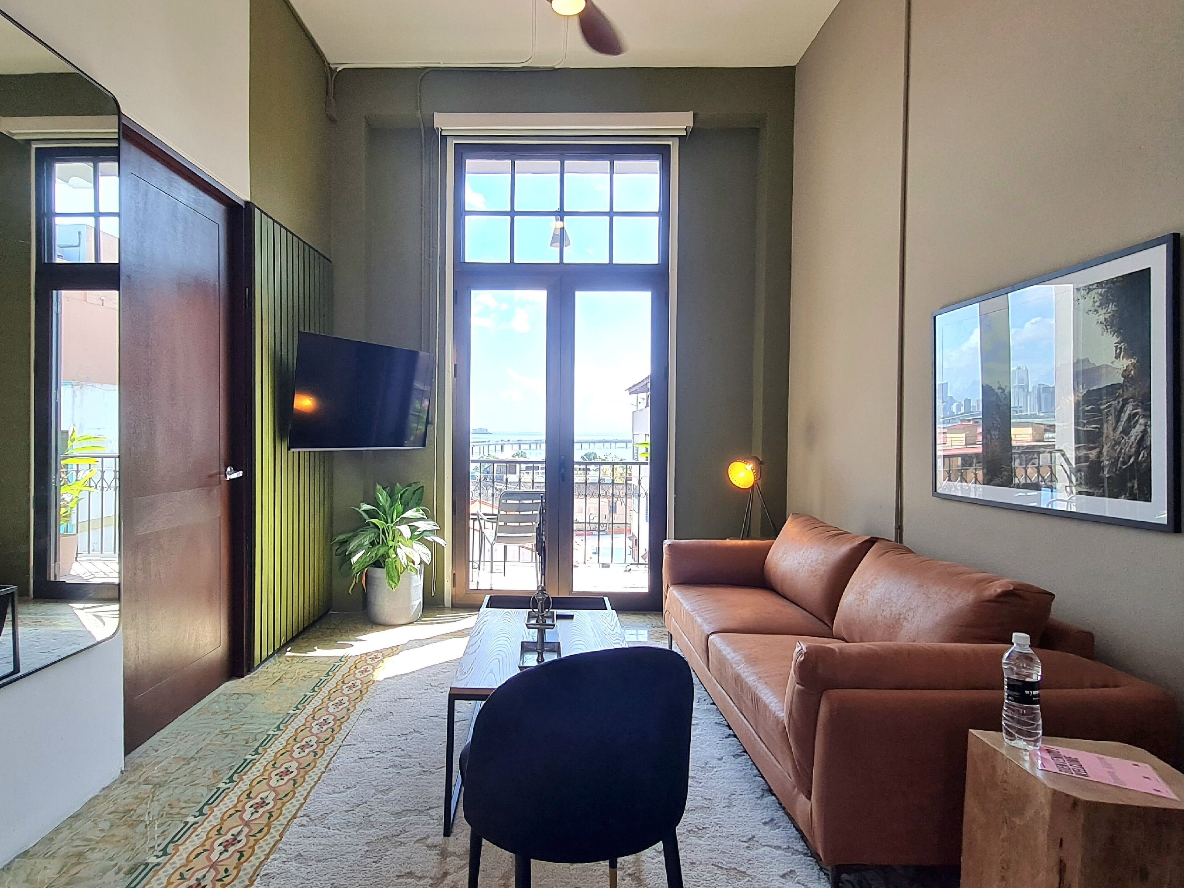 A living room with a brown leather sofa, a black chair, a TV mounted on the wall, a potted plant, and a large window with a view of the outside.