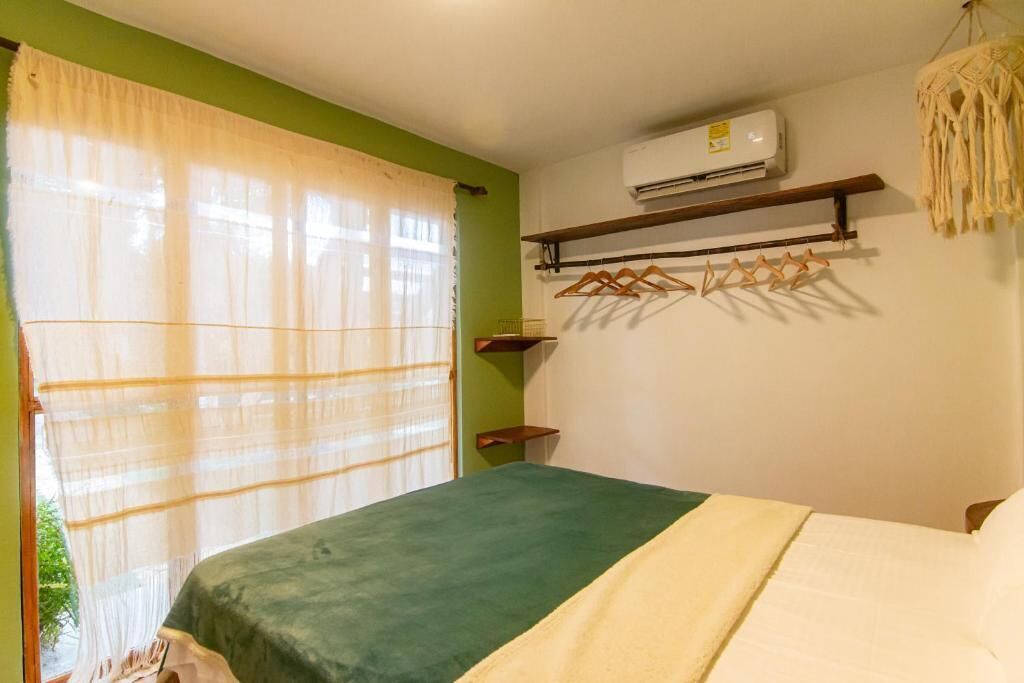 A bedroom with a bed, a window with sheer curtains, an air conditioning unit, and a wall-mounted shelf with a macrame item.