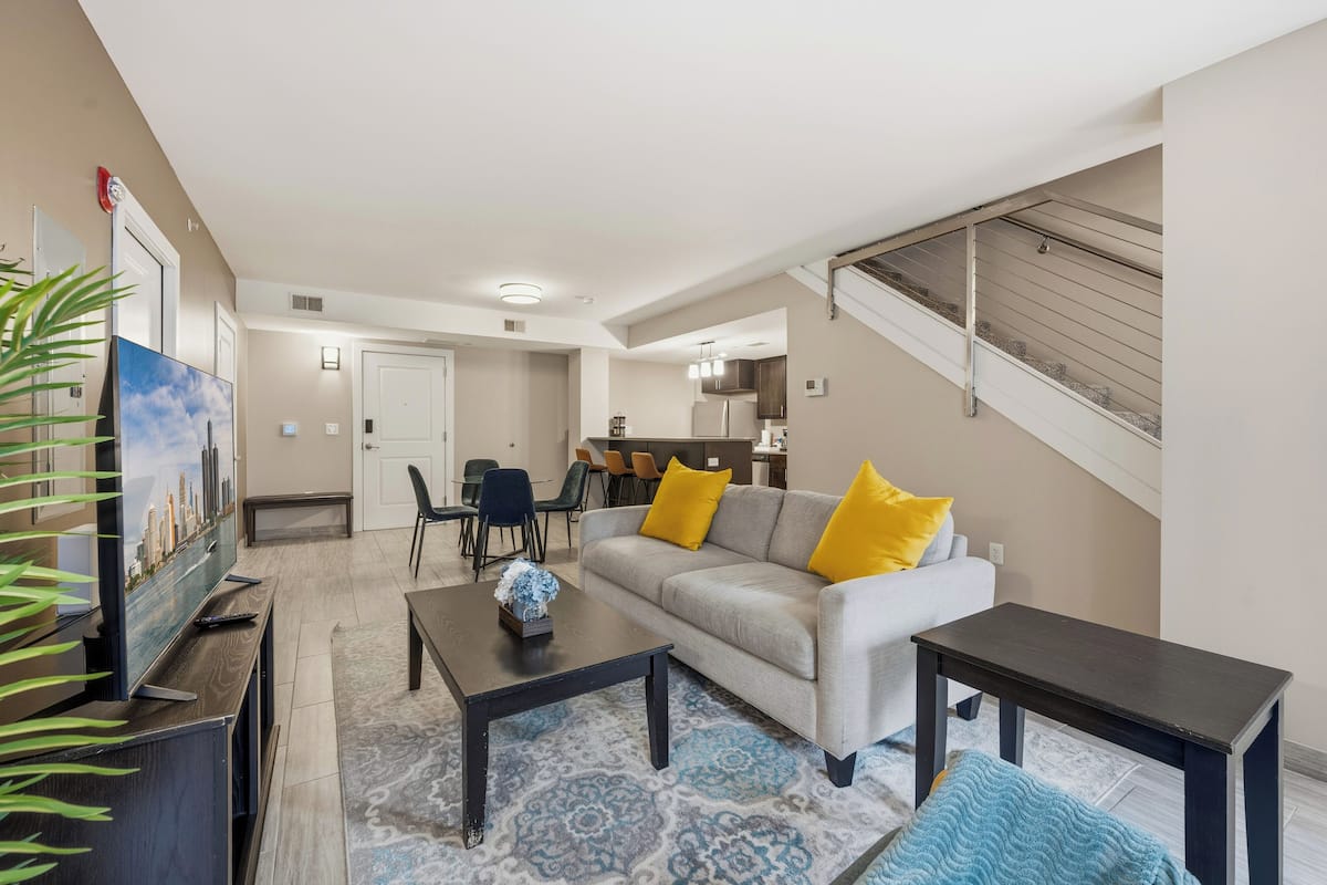 A living room with a sofa, coffee table, and TV stand. There is a staircase leading to an upper level.