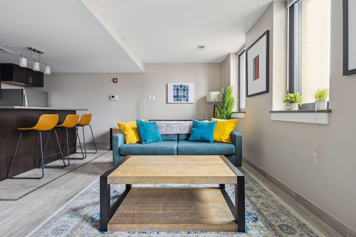 A modern living room with a sofa, coffee table, and chairs, a kitchen area with bar stools, and a window with plants.