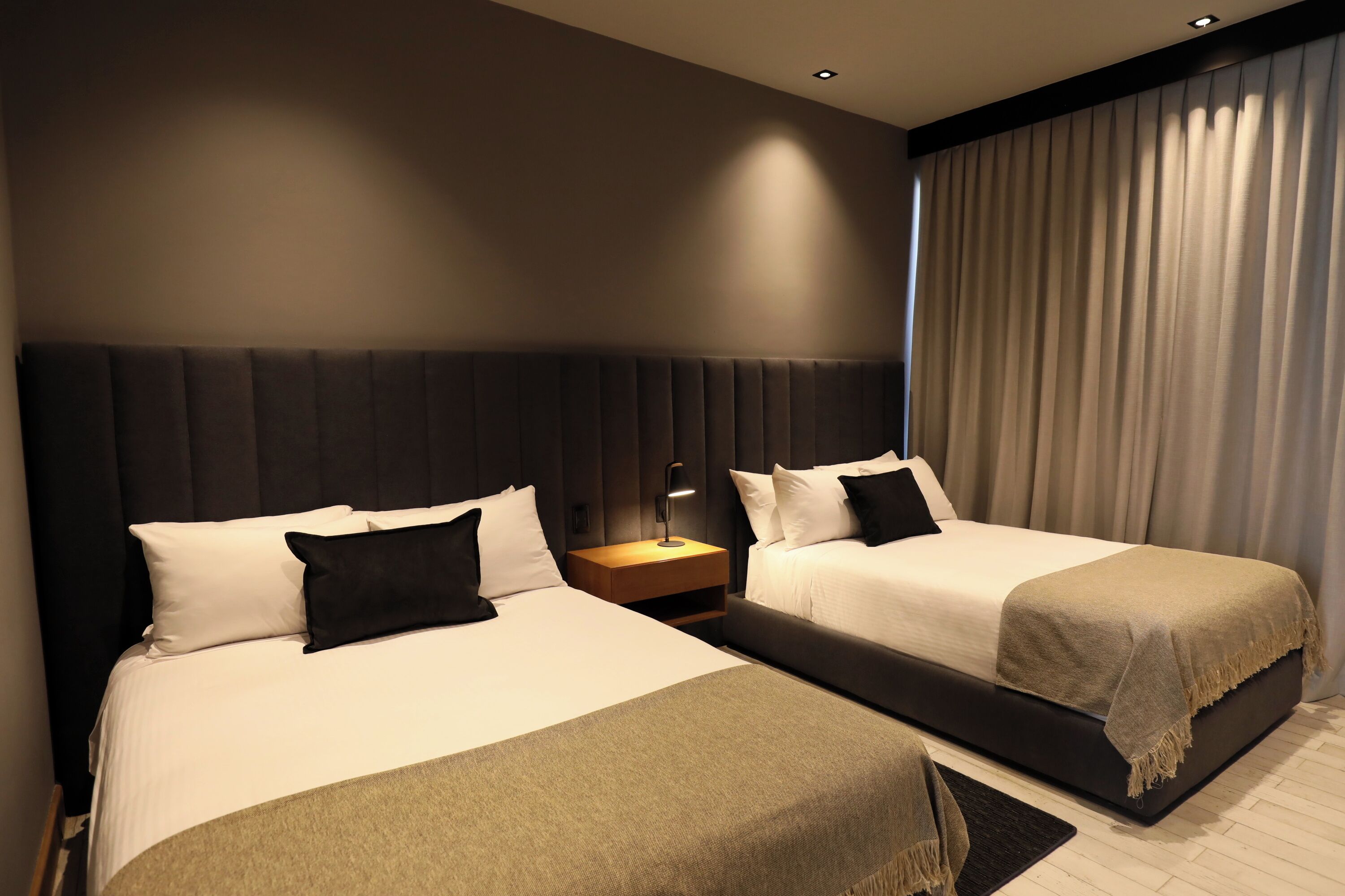 Ein Hotelzimmer mit zwei Betten, einem Nachttisch, einer Lampe und einem großen Fenster mit Vorhängen.