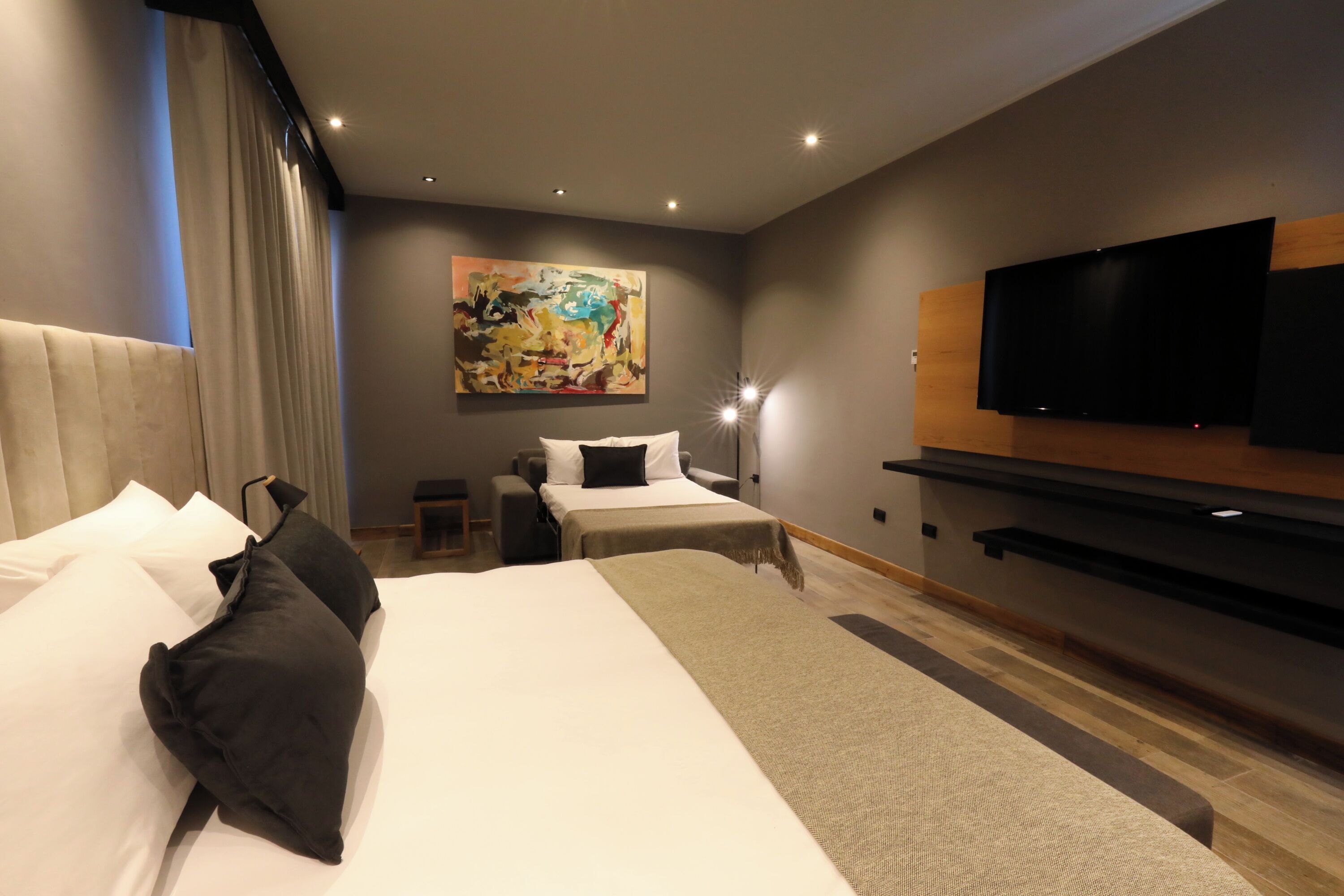 Habitació d'hotel moderna amb un llit gran, un altre més petit, televisió de pantalla plana penjada a la paret i un quadre sobre el llit més petit.