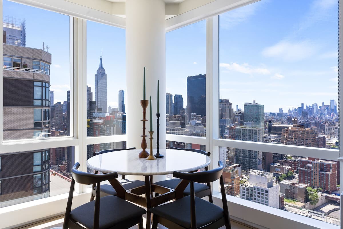 A high-rise room with a large window offering a cityscape view, a round table with chairs, and a decorative vase.