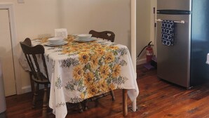 Un comedor con una mesa lista para cuatro personas, una silla de madera, un refrigerador y un extintor.