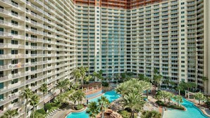 A high-rise hotel with multiple balconies, a central swimming pool, and surrounding palm trees.
