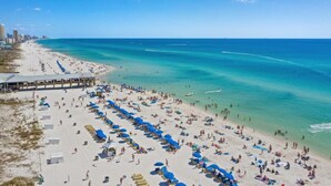 A beach with rows of sun loungers and umbrellas, a pier, and a clear blue sea.
