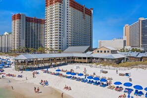 Um resort à beira-mar com vários edifícios altos, um calçadão com guarda-sóis de praia e uma extensa área de areia.