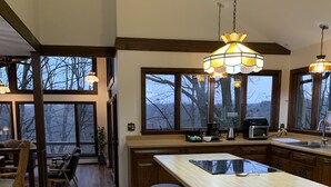 A spacious kitchen with a large island, bar stools, and a view of trees through large windows.