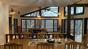 A dining area with a large wooden table, multiple chairs, and a view of the outdoors through large windows.