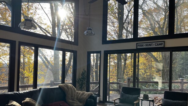 A spacious living room with large windows, a leather sofa, a coffee table, and a sign reading "FISH HUNT CAMP".