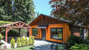 A modern wooden cabin with a covered patio, a wooden pergola, and a well-maintained garden with various plants and a gravel path.