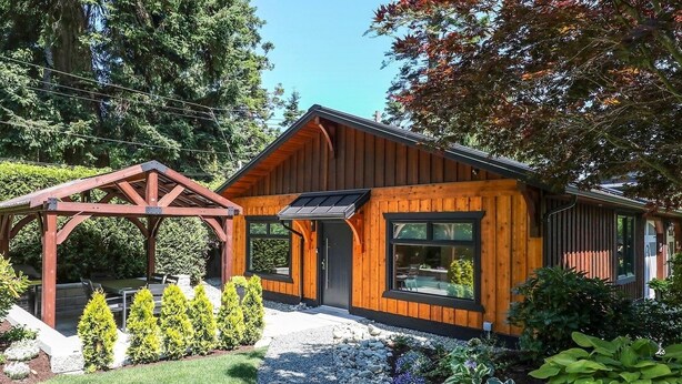 A modern wooden cabin with a covered patio, a wooden pergola, and a well-maintained garden with various plants and a gravel path. - Modern 2BR oceanfront apartment for sun, ski or mountain biking! (Courtenay)