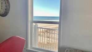 A room with a large window overlooking a beach, a clock on the wall, and a red chair. - Sunchase #303: Steps to Beach! Great View! (Gulf Shores)