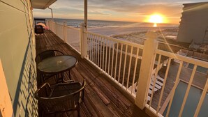 A wooden deck with a glass table and chairs, overlooking a beach with a sunset. - Sunchase #303: Steps to Beach! Great View! (Gulf Shores)