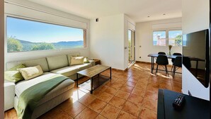 Una sala de estar moderna con sofá, mesa de centro y televisor. Hay un comedor con sillas y una mesa. Amplias ventanas ofrecen vistas al exterior.