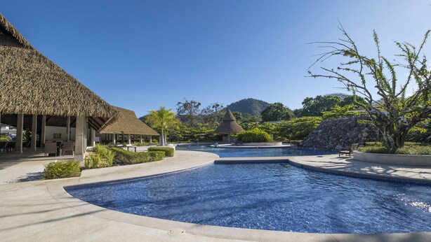 A resort pool area with a thatched-roof building, a small hut, and a mountain backdrop.