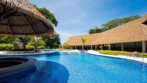 A swimming pool with a thatched roof structure, surrounded by lounge chairs and greenery. - Casa Celeste lovely beach house (Jacó)