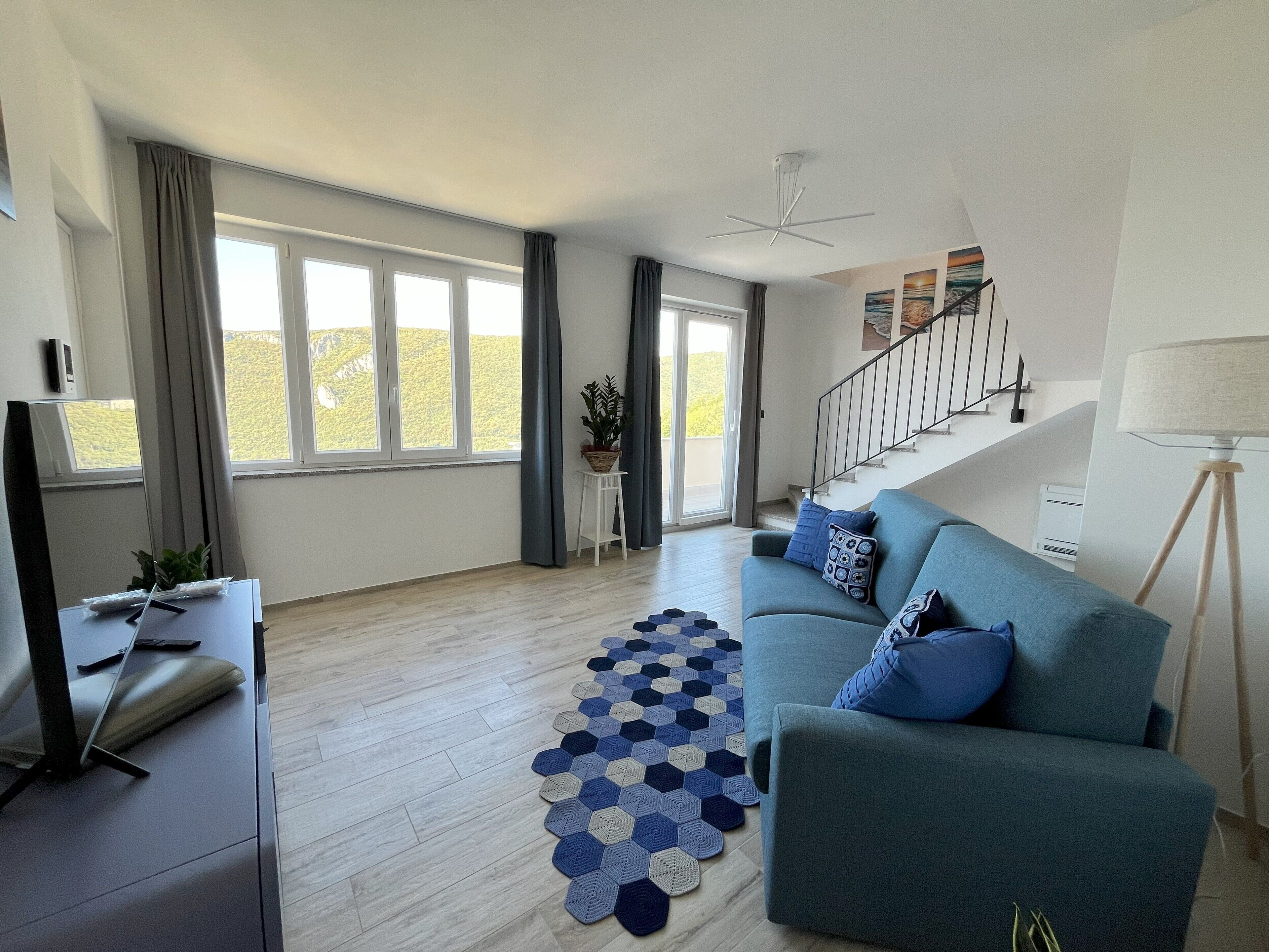 A modern living room with a sofa, a coffee table, and a TV. There is a staircase leading to another level, and a large window with curtains.