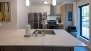 Une cuisine moderne dotée d’un îlot central, d’appareils électroménagers en acier inoxydable, de luminaires suspendus et offrant une vue sur l’extérieur.