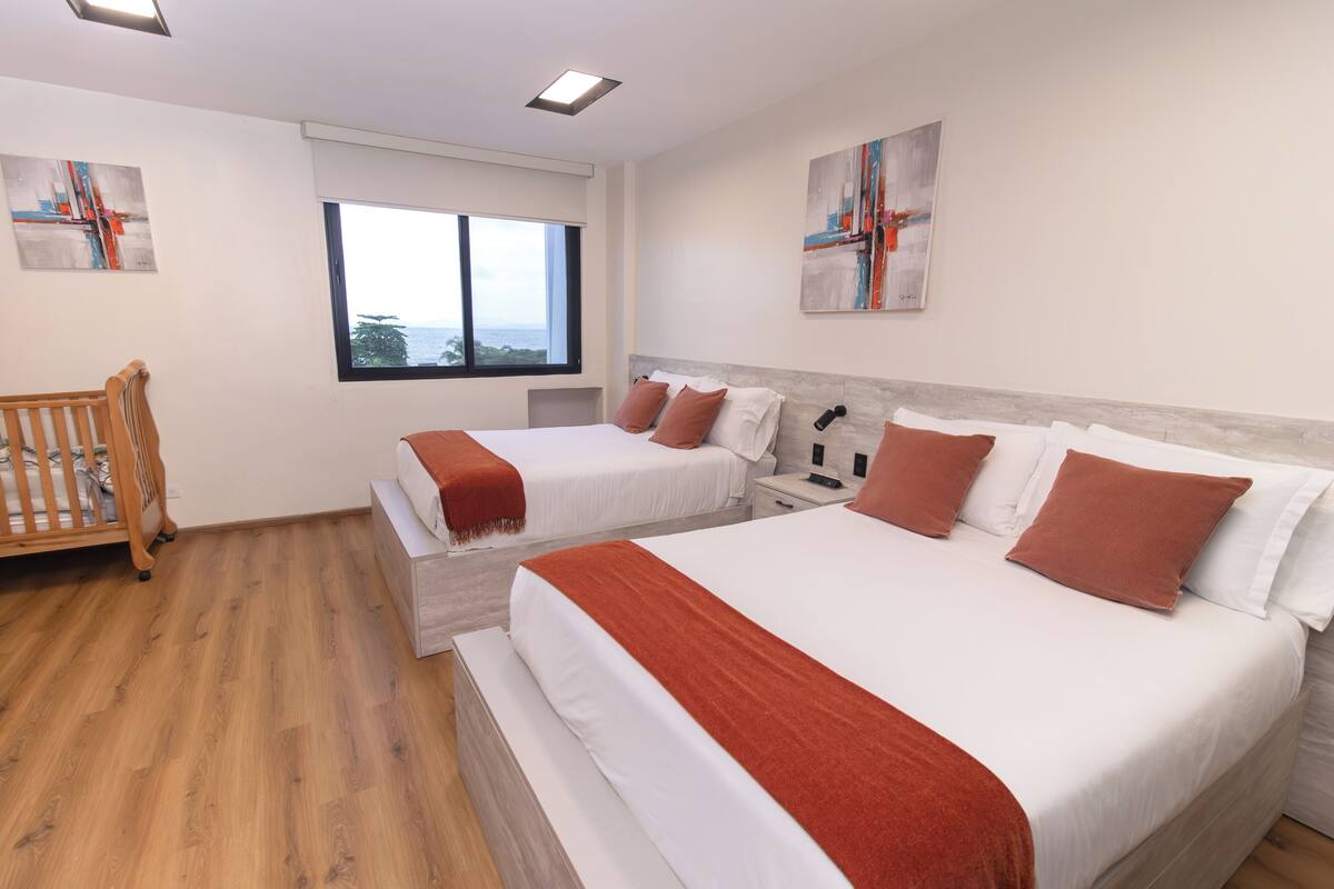 A hotel room with two beds, a crib, a large window, and artwork on the walls.