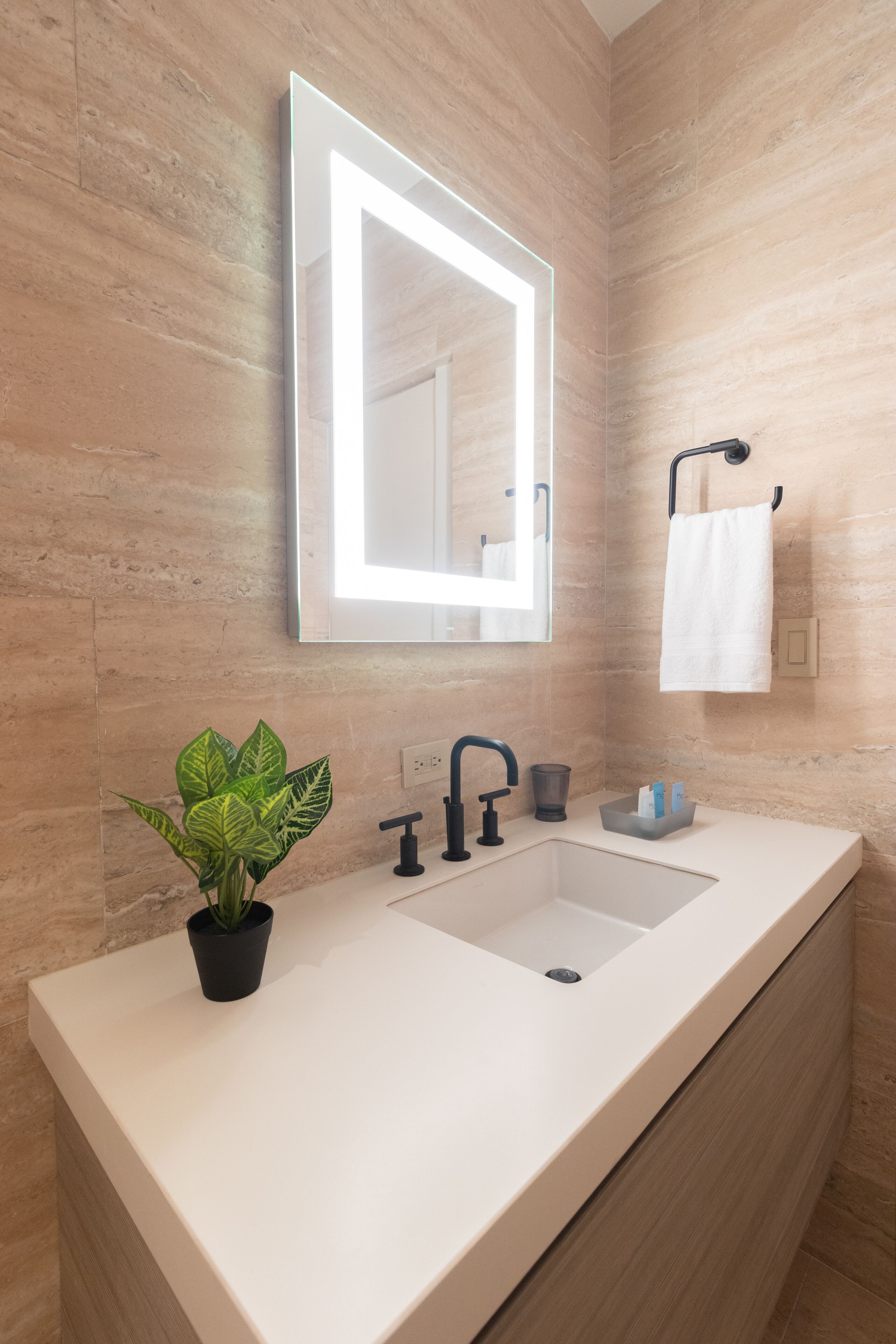 Un bagno moderno con un lavandino bianco, uno specchio con illuminazione, un porta asciugamani e una pianta in vaso.