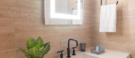 Un bagno moderno con un lavandino bianco, uno specchio con illuminazione, un porta asciugamani e una pianta in vaso.