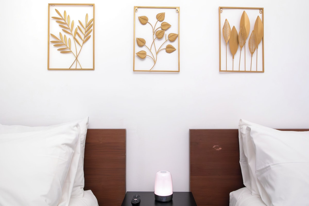 Een hotelkamer met twee bedden, een nachtkastje met een afstandsbediening en een klok, en drie ingelijste kunstwerken aan de muur.