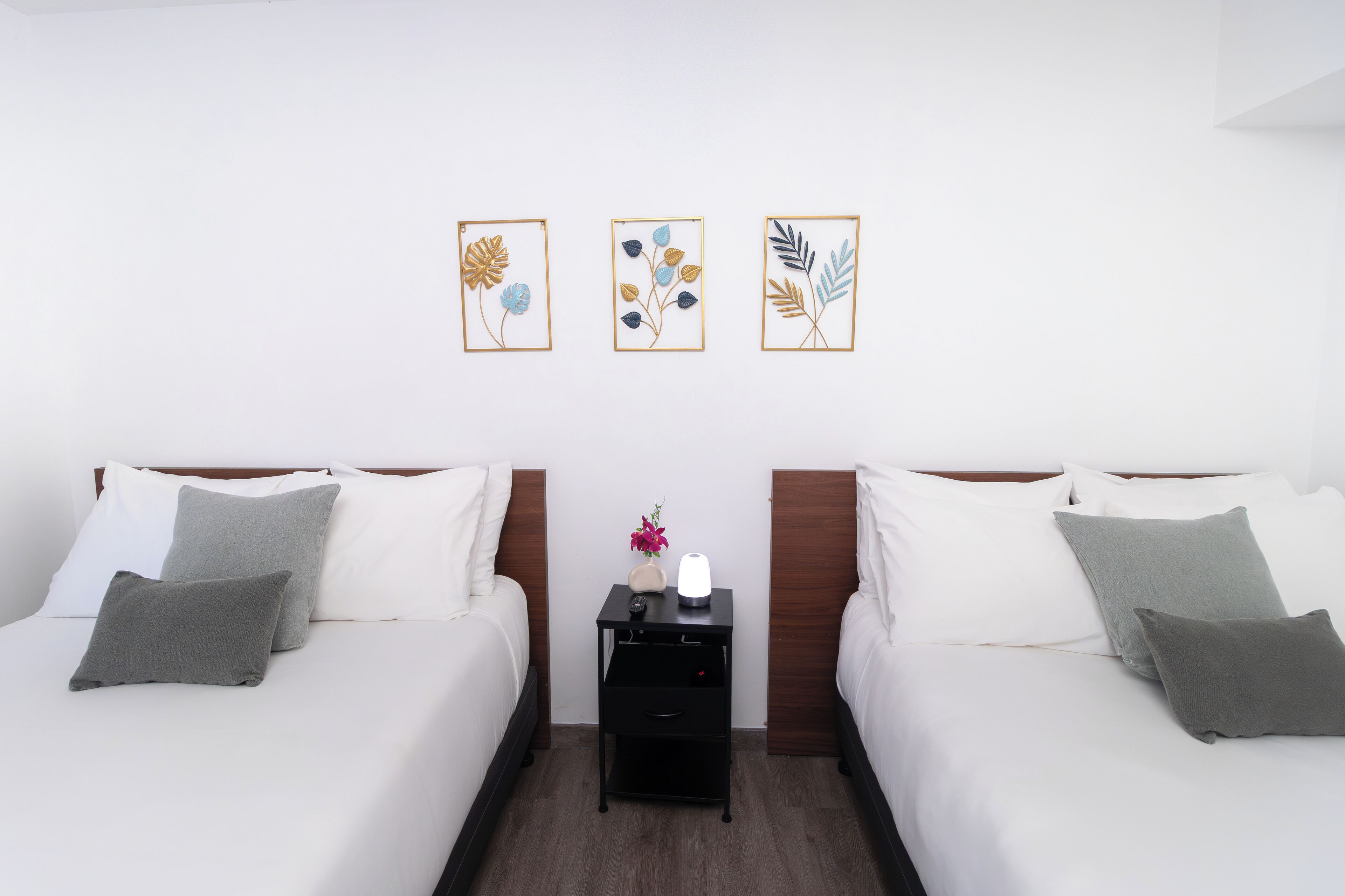 Une chambre d’hôtel avec deux lits, une table de chevet avec une lampe et trois œuvres d’art florales encadrées accrochées au mur.