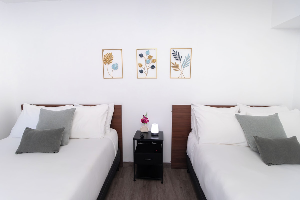 A hotel room with two beds, a nightstand with a lamp, and three framed floral artworks on the wall.