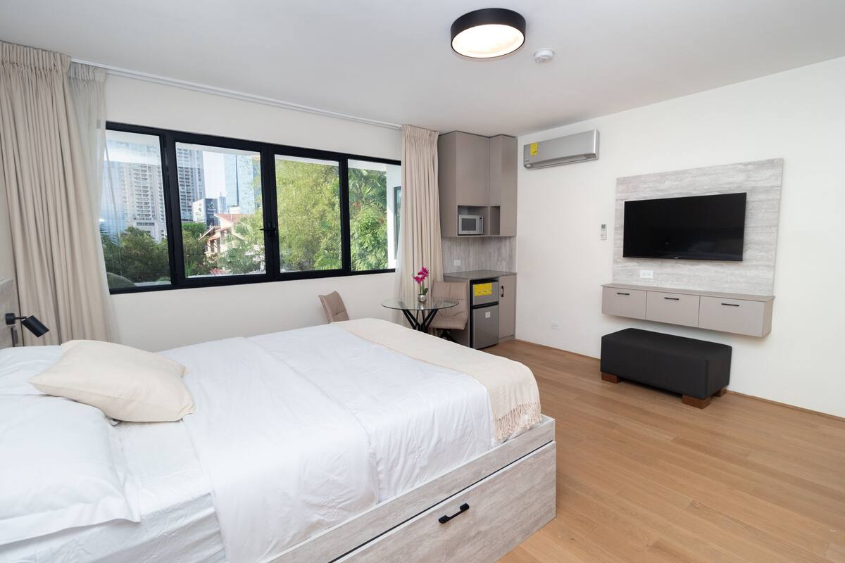 A modern bedroom with a large bed, a TV mounted on the wall, a small kitchenette, and a view of the city through large windows.