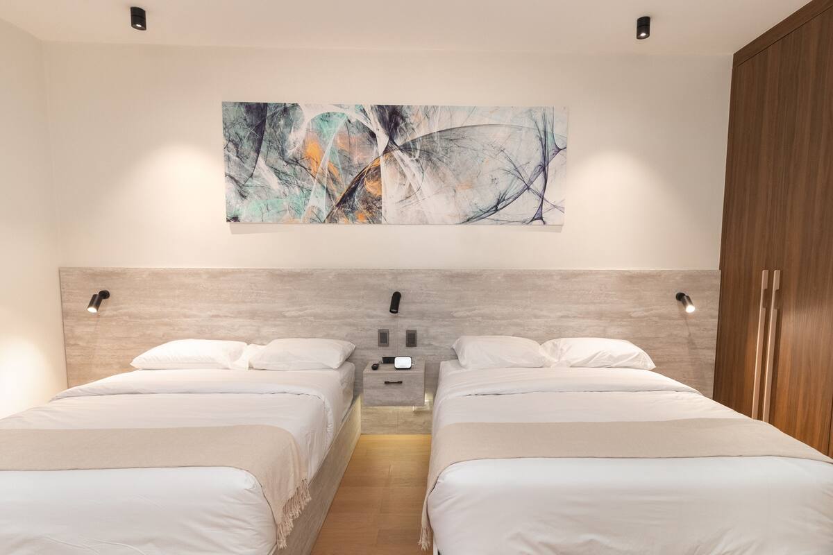 A modern hotel room with two beds, a large abstract painting above the headboard, and a wooden wardrobe.