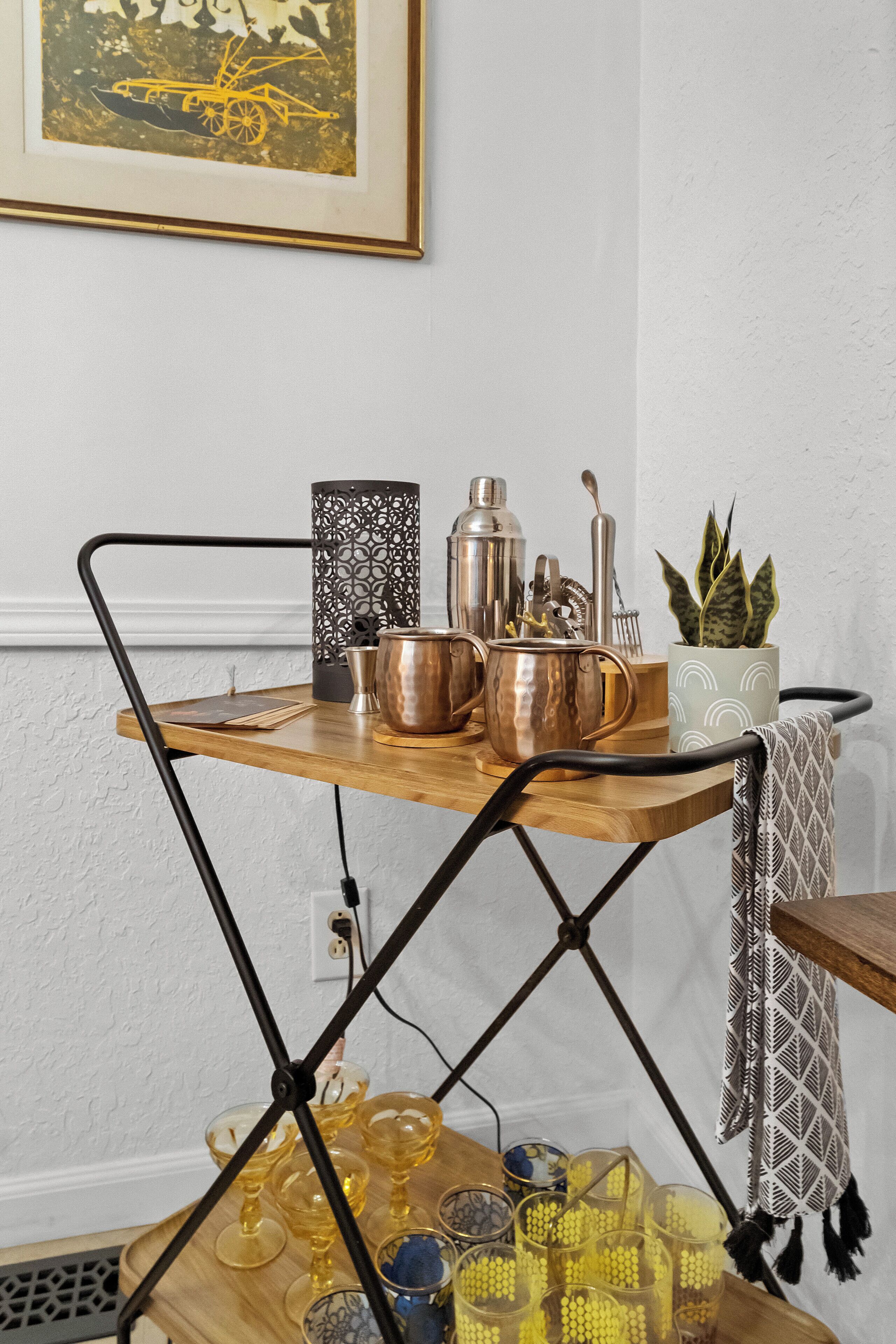 Un carrito de bar de madera con detalles metálicos, una planta en maceta y diverso utensilio de cocina.