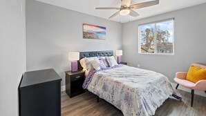 Une chambre à coucher comprenant un lit, des tables de chevet, une chaise, un ventilateur de plafond, une fenêtre et un tableau accroché au mur.