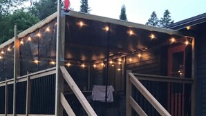 A wooden deck with a staircase leading to a covered area, string lights, and a small table with a chair. - Cozy Cabin In The Woods  (Sprucedale)