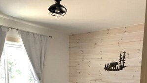 A bedroom with a bed, a nightstand, a window with curtains, and a wall decal of a bear and trees. - Cozy Cabin In The Woods  (Sprucedale)