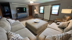 Een woonkamer met een beige hoekbank, een salontafel, een televisie op een houten dressoir, een plafondventilator en een deur met een raam.