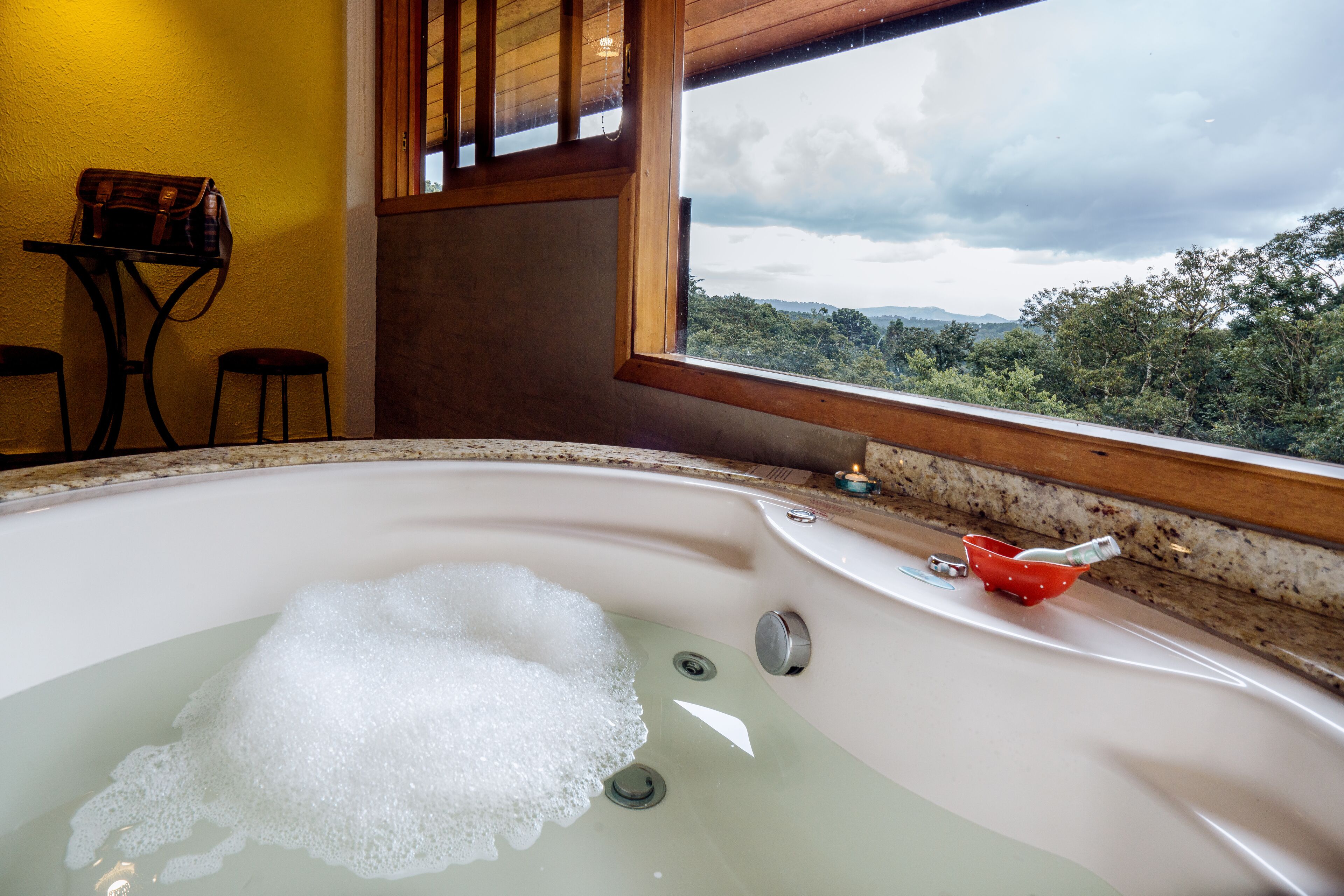 A bathroom with a large bathtub filled with bubbles, a wooden table with a bag, and a view of a forest through a window.