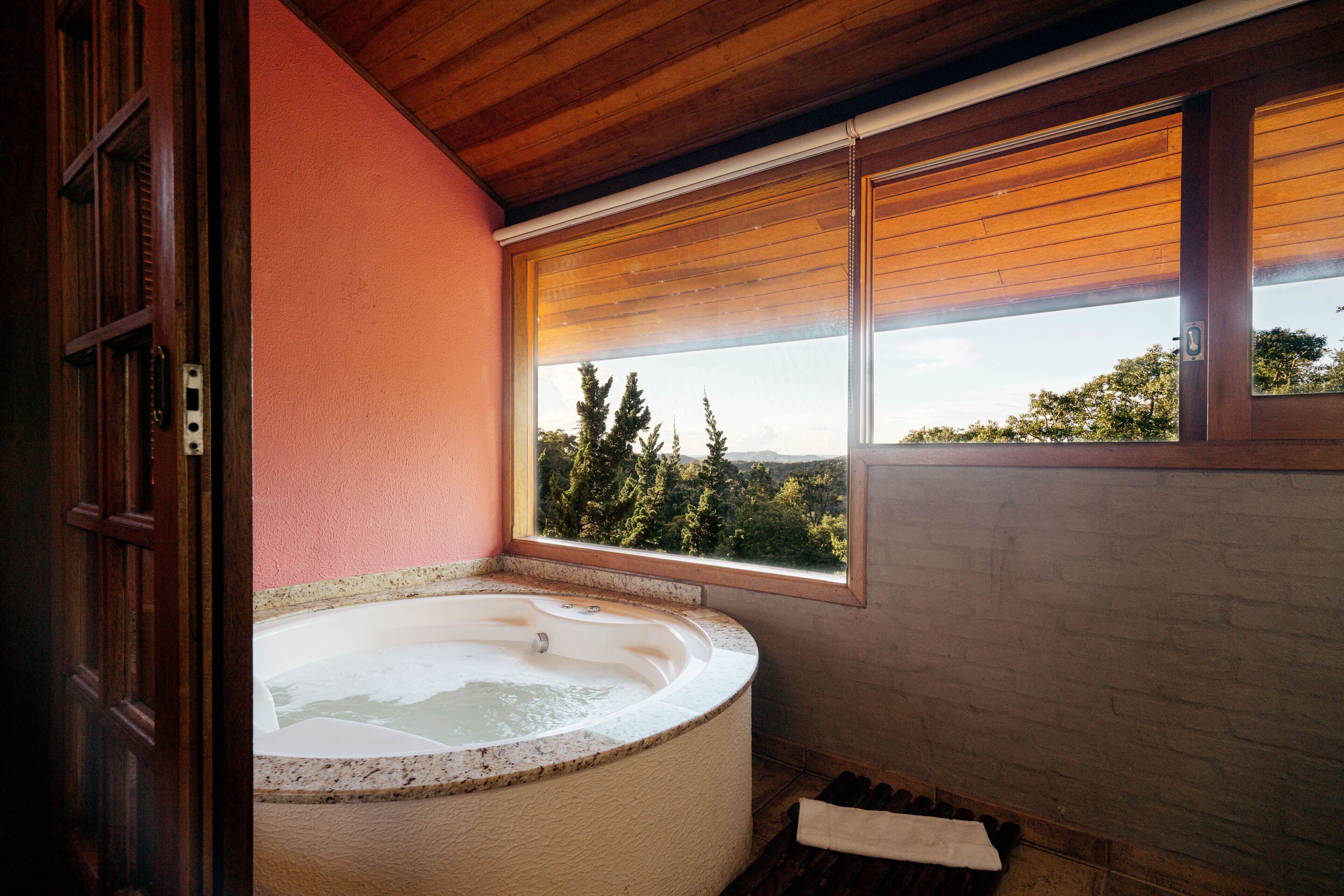 Un baño con una bañera grande, una ventana con persianas y vistas a árboles y montañas.